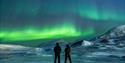 Two people stand on the ice, observing the majestic winter landscape illuminated by the green northern lights in the sky.
