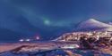 A snowmobile in the foreground with buildings in Longyearbyen, snow-covered mountains and northern lights in the sky in the background.