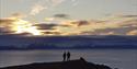 Two persons on top of a mountain in silhouette against a dark fjord and a sunset behind low clouds in the background