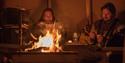 Inside the lavvu. In the foreground, a fire is lit, and in the background, two people are sitting and eating hot food.