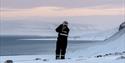 En gjest i snøscooterutstyr som står på en snødekt haug og speider utover en fjord og snødekte landskap i bakgrunnen
