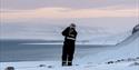 A guest wearing snowmobile equipment standing on a snowy hill scouting out towards a fjord and snowy landscapes in the background