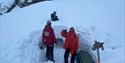 Two people standing at the entrence of a snow cave. One person laying at the stow above the entrence.