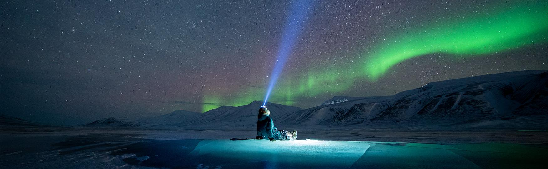 A person sitting on an ice formation with a headlamp on their head while looking up at a clear starry sky with northern lights in the background