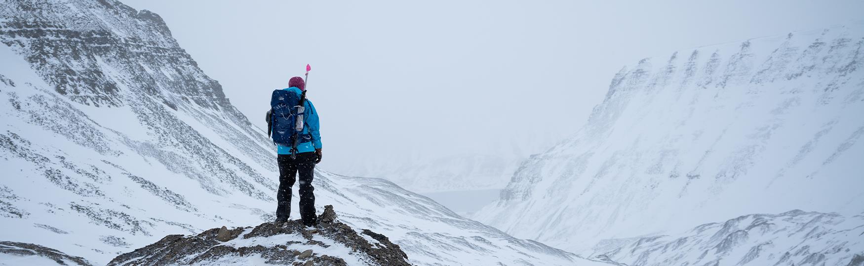 A guide with a backpack and a rifle on top of a cliff looking at a snow-covered mountainous landscape