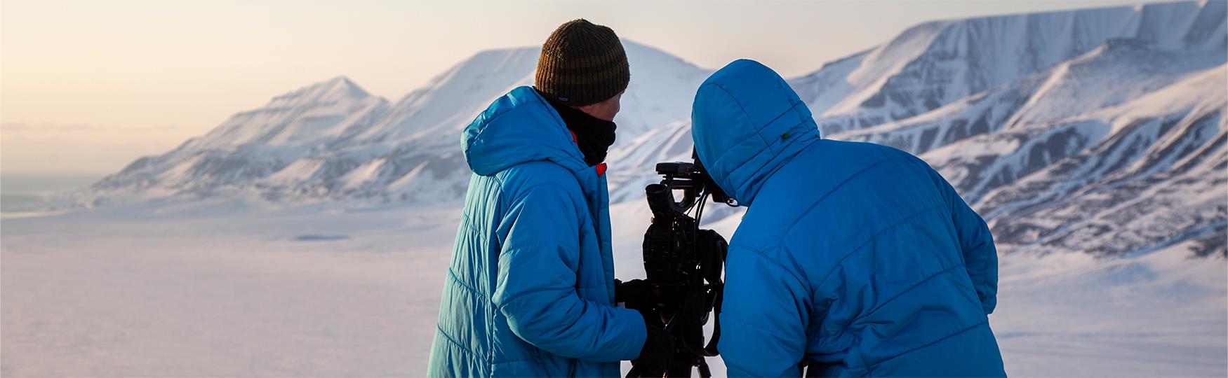 Two persons looking at a camera on a tripod with snow-covered mountains in the background