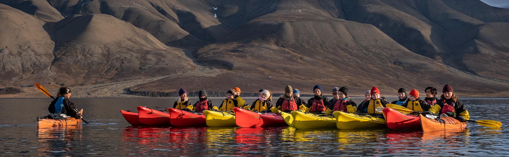 A group of guests and a guide in kayaks on a fjord with mountains in the background