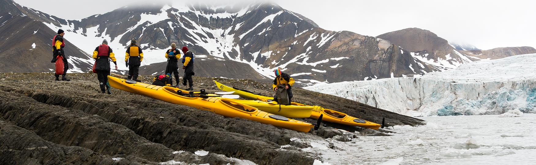 A group of guests and a guide in kayaking equipment next to several kayaks on the shore of a fjord. In the background there's a large mountain and a glacier.