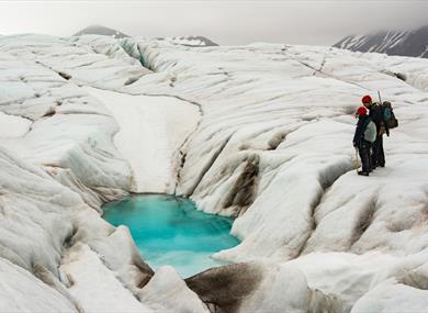 Two persons standing on a glacier, looking down on a meltingwatercanal 