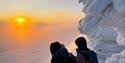 Tre personer står på toppen av fjellet, gjennom et tynt lag med tåke kan man se en tydelig solnedgang.