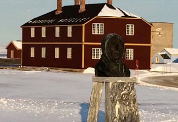 En byste av Roald Amundsen i metall i sentrum av Ny-Ålesund, med bygninger og snødekte gater i bakgrunnen