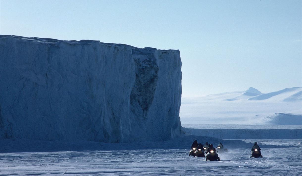 En turgruppe på snøscootere med en isvegg i bakgrunnen