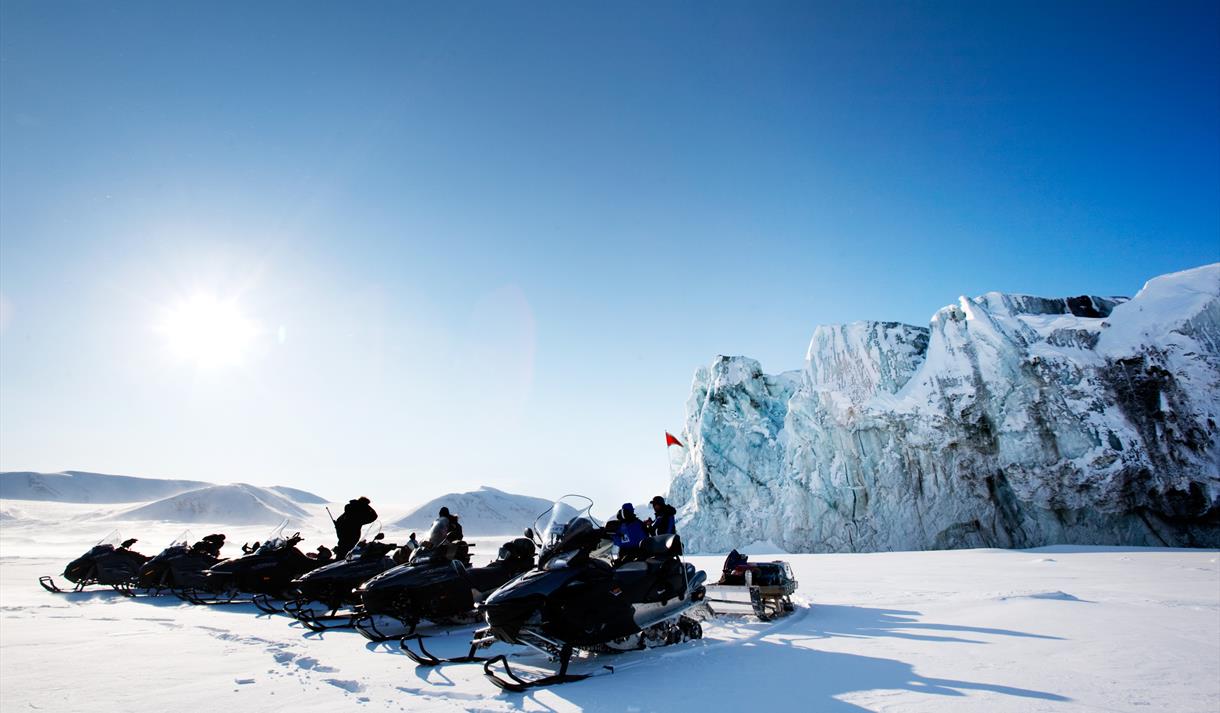 En rekke med snøscootere på en snødekt slette med en høy isbrefront og en blå himmel i bakgrunnen