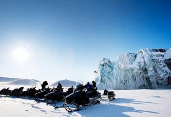A line of snowmobiles on a snowy plain with a tall glacial front and a clear blue sky in the background