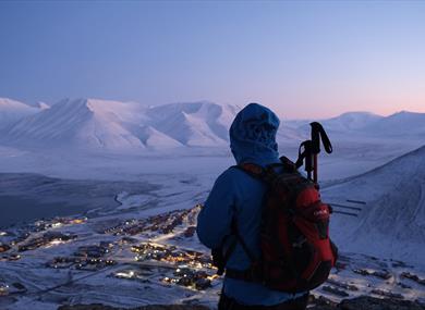 A person looking out across a snow-covered landscape from a mountain with Longyearbyen situated below and a mountainous landscape in the background