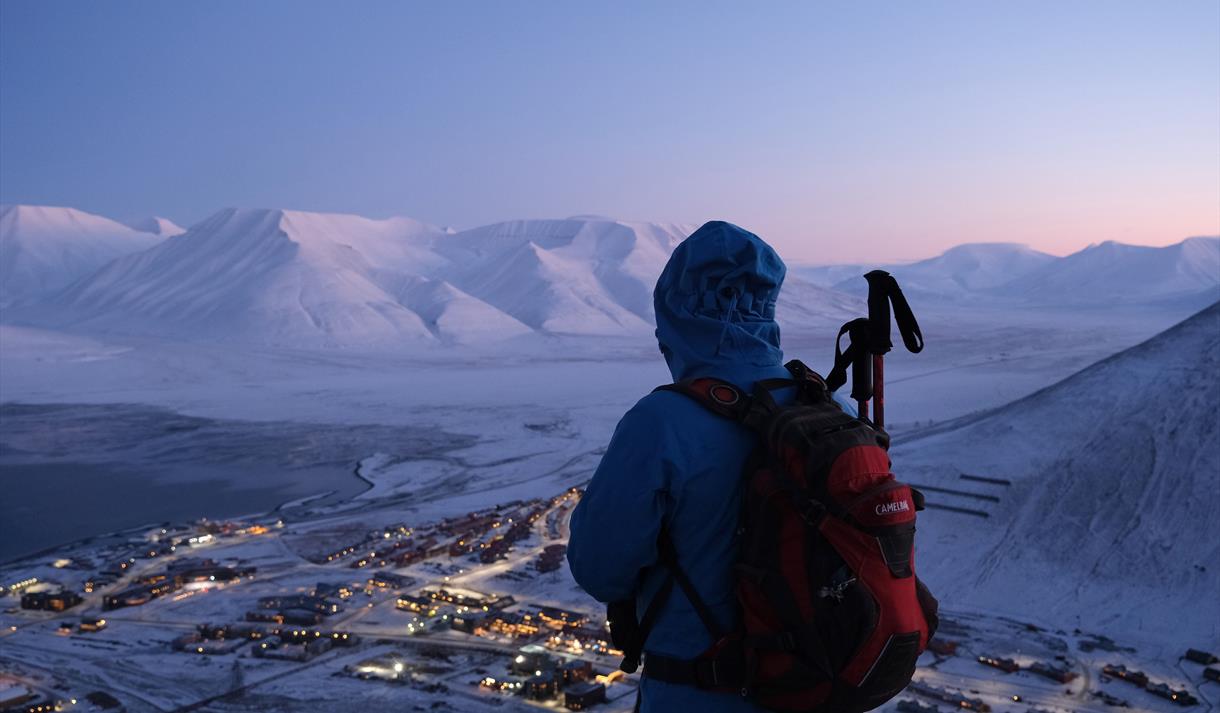 A person looking out across a snow-covered landscape from a mountain with Longyearbyen situated below and a mountainous landscape in the background