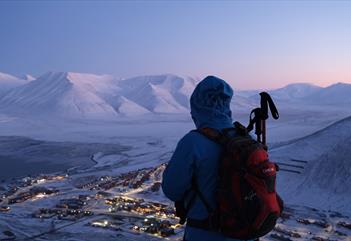 En person som ser ut over et snødekt landskap fra et fjell med Longyearbyen nedenfor seg selv og et fjellandskap i bakgrunnen