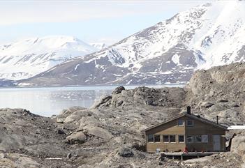 The wilderness cabin Nordenskiöld Lodge surrounded by a rocky morraine landscape in the foreground, with an open fjord and partially snow-covered moun