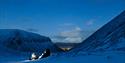 To personer med hodelykt i forgrunnen, med fjell og Longyearbyen i bakgrunnen.