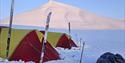 To telt og en pulk i en teltleir i forgrunnen, med et åpent snødekt landskap og et spisst fjell i bakgrunnen