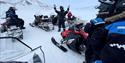 Several people with snowmobiles, having a break. One person standing with the hands in the air, telling something to the rest.