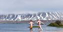 To personer som løper ut i Isfjorden for å bade. I bakgrunnen ligger det fjell med snø på.