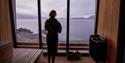 En person som står i badstuen på Isfjord Radio iført en badekåpe og ser ut over Isfjorden gjennom et vindu.