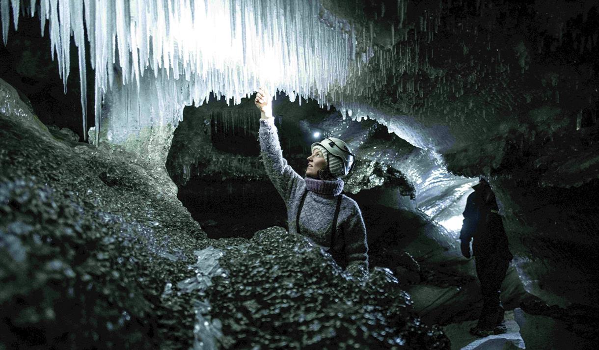 En guide med en lys hodelykt og hjelm på inne i en isgrotte som kjenner på en istapp i taket av grotten