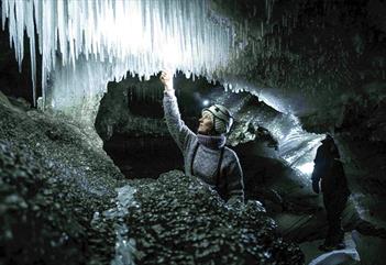 A guide with a bright head lamp and helmet inside an ice cave feeling one of the icicles hanging from the cave roof