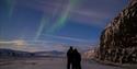 Two persons in snowmobile equipment hugging with northern lights in the skies above
