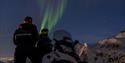 Two persons standing behind a snowmobile and looking up at northern lights in the sky