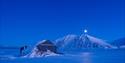 Et snødekt landskap i blått lys med en nedsnødd hytte i forgrunnen