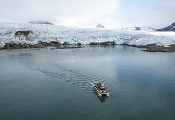 En båt som seiler på en fjord i forgrunnen med en isbrefront og fjell rundt en bukt i bakgrunnen