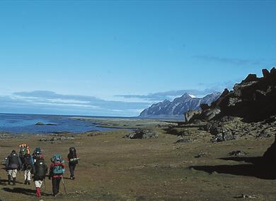 A group of guests and a guide hiking with large backpacks in the foreground along a seafront with tall mountains and a blue sky in the background