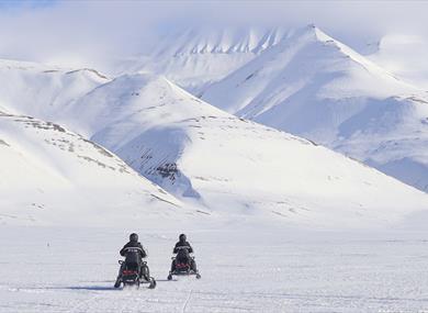 Two persons on electric snowmobiles in the foreground driving across a frozen snowy plain towards snow-covered mountains in the background