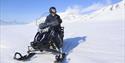 A smiling person wearing snowmobile equipment driving an electrical snowmobile through a snow-covered landscape