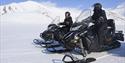 Two persons in snowmobile equipment on snowmobiles smiling to each other, surrounded by a snowy landscape
