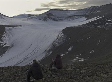 Two guests sitting in a rocky area looking towards the glacier Foxfonna in the background