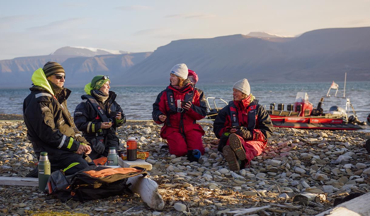 Two guides and two guests sitting on a beach talking with each other, and a RIB boat anchored by the shore of the fjord in the background