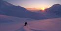 A person skiing down a mountain at dusk with a sunset over a mountain ridge in the background