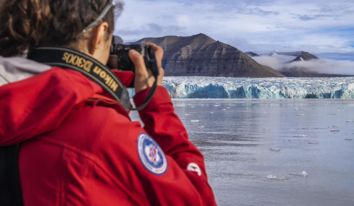 A person with a camera in the foreground taking photos across a fjord of a blue glacier front and mountains in the background