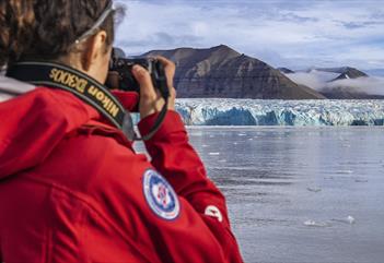 A person with a camera in the foreground taking photos across a fjord of a blue glacier front and mountains in the background