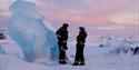To gjester i snøscooterutstyr som står ved siden av et stort blått isfjell på sjøis og prater med hverandre
