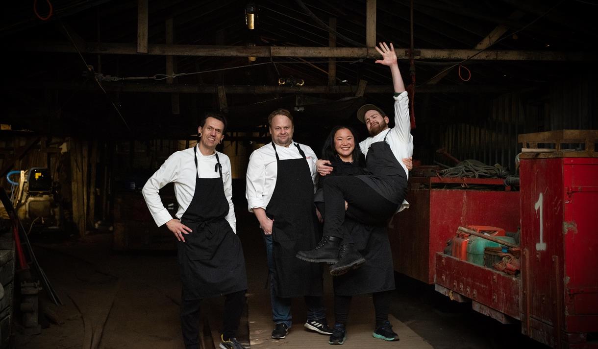 Four persons in chef's clothes standing in an industry hall. A person to the right is lifting another with their arm outstreched into the air.
