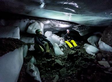 Two people sitting inside an ice cave, looking at the formations on the roof.