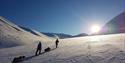 Two persons skiing while pulling sleds on their way towards the sun in a snowy landscape