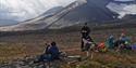 Two people sitting down to rest together with a pack of dogs during a hike, with a bare tundra landscape and a glacier up in the mountains