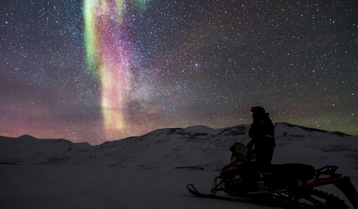 En person står oppreist på en parkert snøscooter og ser på den klare himmelen som bærer preg av tydelig nordlys og stjerner.