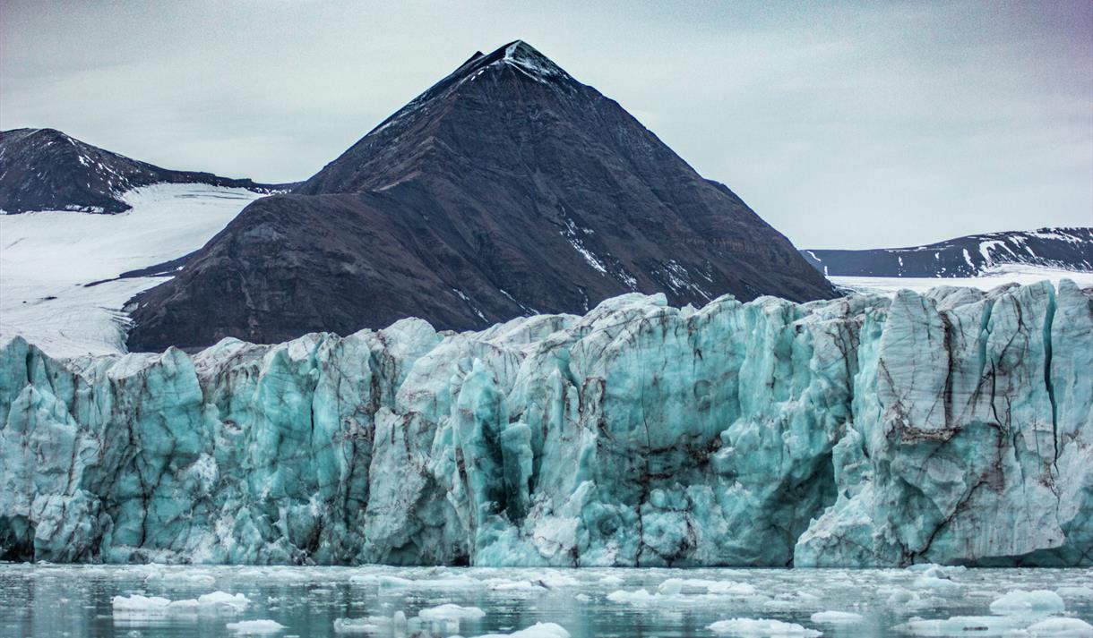 En fjord med flytende isbiter og en brefront som stiger opp mot fjell i bakgrunnen