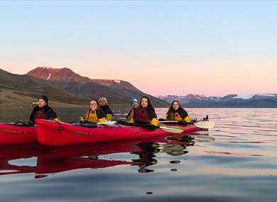 A group of guests in kayaks on a calm fjord with mountains in the background